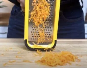 Grated carrots on a wooden board using a bright yellow box grater in a kitchen setting.